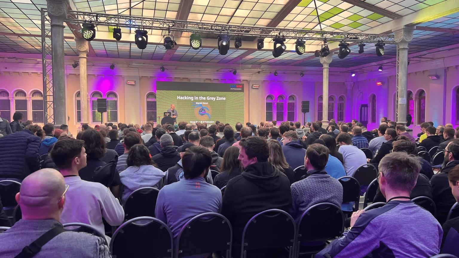 Large audience at cybersecurity conference BSides Prague listening to presentation titled "Hacking in the Grey Zone" in ornate venue with purple lighting and professional stage setup