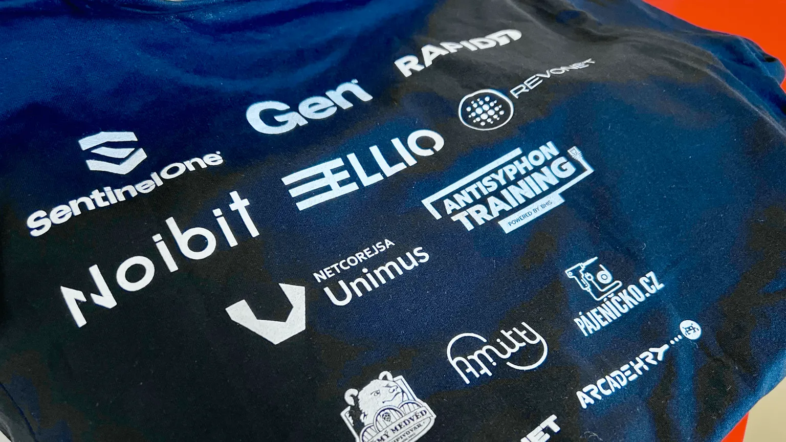 BSides Prague T-Shirt displaying various cybersecurity company logos including SentinelOne, Noibit, Gen, Rapid7, ELLIO, Antisyphon Training, Netcore ISA Unimus, Amity, Pauenino.cz, and ArcadePro