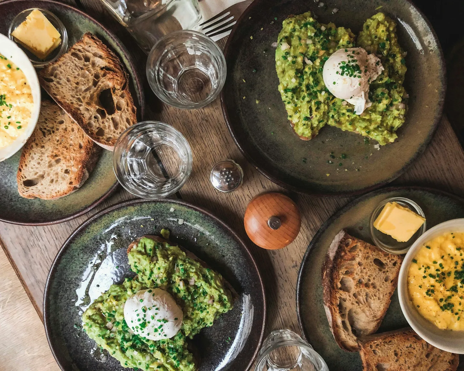 Overhead view of breakfast table with avocado toast topped with poached eggs, sourdough bread, butter, scrambled eggs, and water glasses on dark ceramic plates